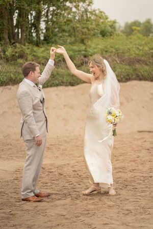Bride and groom dancing joyfully on sandy beach on their wedding day.
