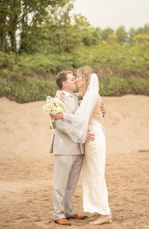 Bride and groom share a kiss on sandy beach, bride in white gown with veil holding yellow bouquet, groom in tan suit.