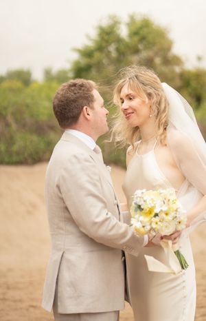 Smiling bride in white gown holds bouquet while gazing at groom in beige suit outdoors.