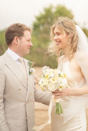 Smiling bride in white gown holding yellow bouquet gazes at groom in beige suit outdoors.