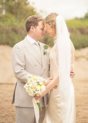 Bride and groom touching foreheads tenderly on a sandy beach holding a white floral bouquet.