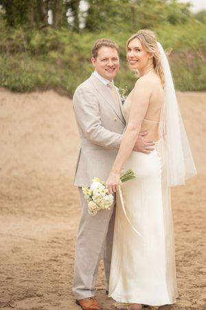 Bride in backless white halter gown and veil stands with groom in beige suit on a sandy beach holding bouquet.