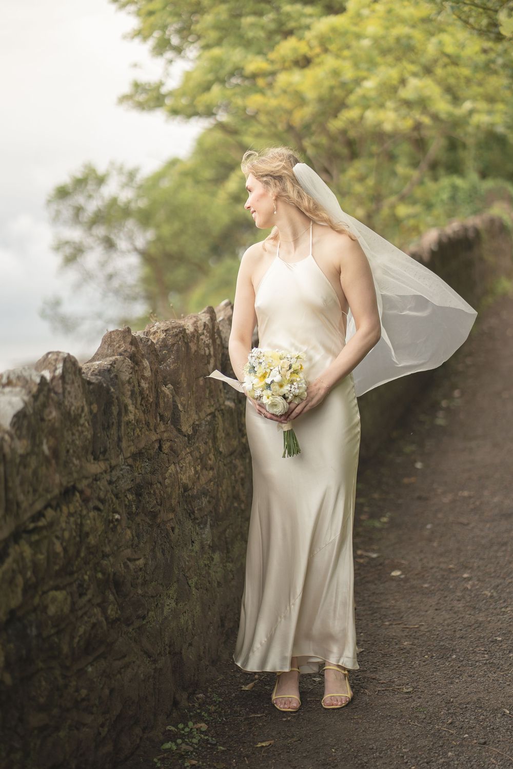 Elegant bride in satin gown holding bouquet beside stone wall outdoors.
