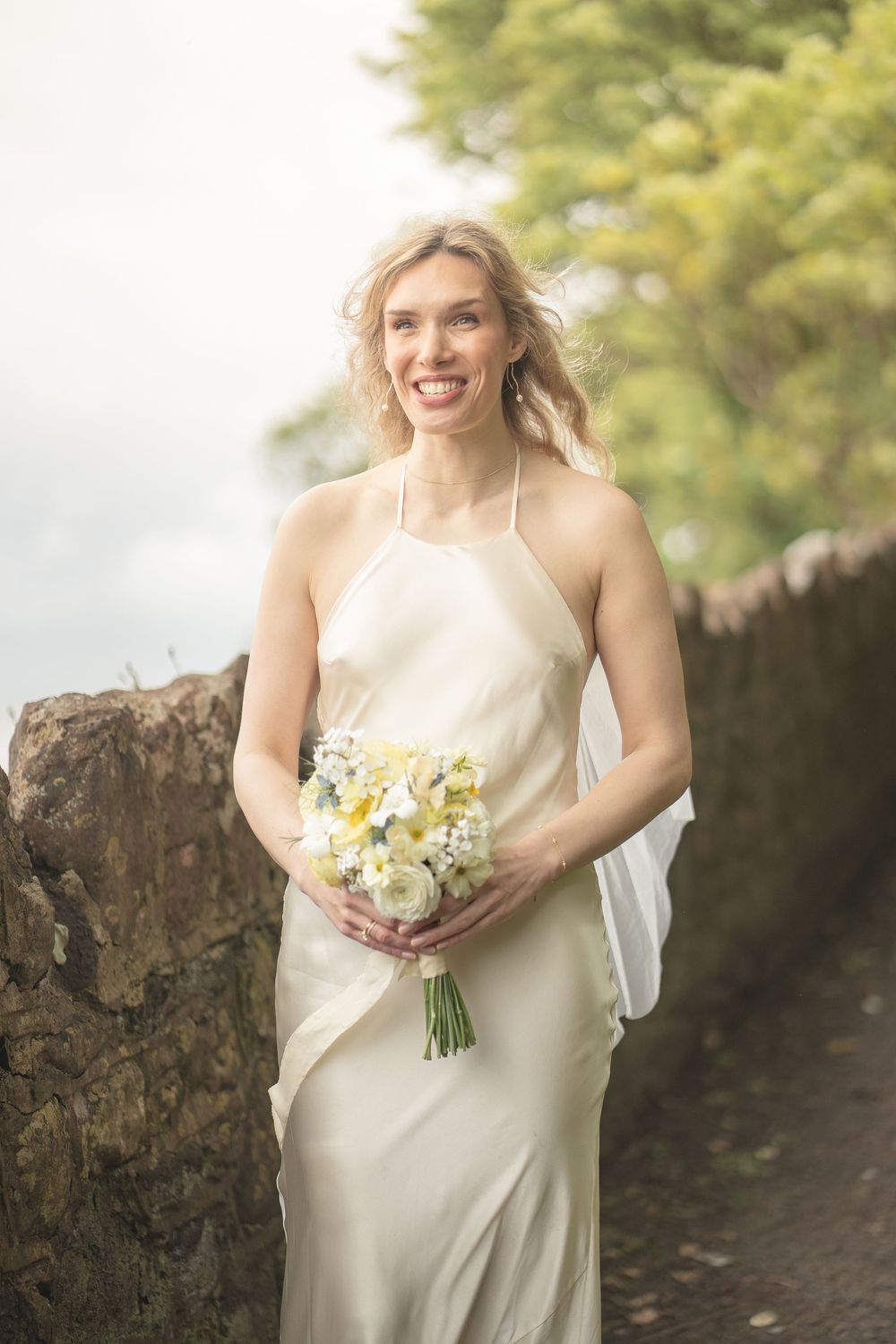 Smiling bride in ivory halter gown holding white and yellow bouquet outdoors near stone wall.