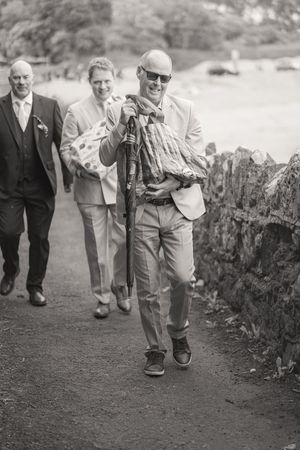 Black and white photo of smiling bald man in sunglasses carrying umbrella and bags on a path with two men behind him.