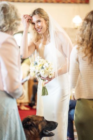 Smiling bride in white halter gown and veil holds yellow and white bouquet while guests and a dog gather around her.