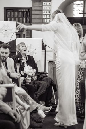 Black and white photo of a bride in a white gown gesturing to laughing wedding guests seated in church pews.