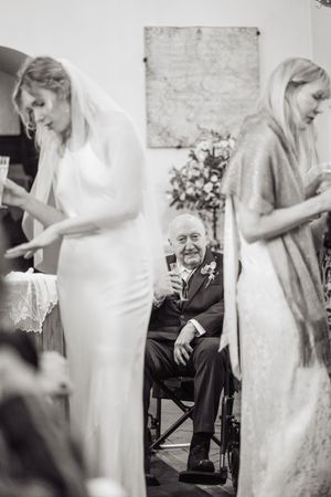 Elderly man in suit raises champagne glass between bride in veil and blonde woman at wedding ceremony.