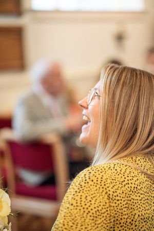 Smiling blonde woman with glasses in a yellow polka dot dress laughing at a wedding ceremony indoors.