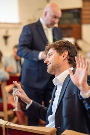A curly-haired man holds a champagne glass and gestures while seated in a church pew.