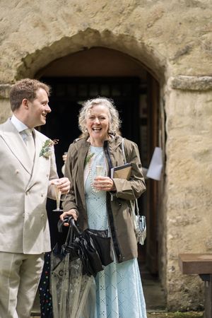 A laughing woman in a blue dress and olive jacket holds champagne with a groom in a tan suit outside a stone church.