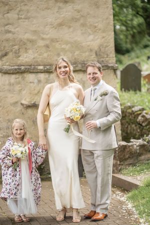 Bride and groom posing with young flower girl outside a stone church holding yellow bouquets.