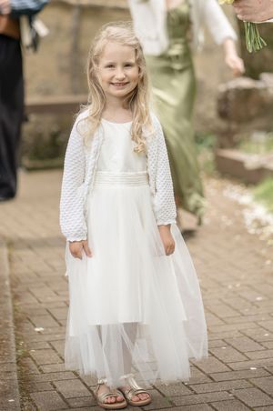 Smiling flower girl in white tulle dress and knit bolero jacket at a wedding ceremony.