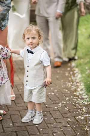Adorable toddler page boy in beige vest and bow tie standing on a brick pathway at a wedding.