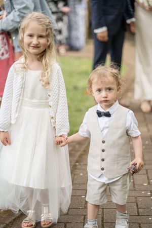 Flower girl in white dress and page boy in beige vest holding hands at a wedding ceremony.