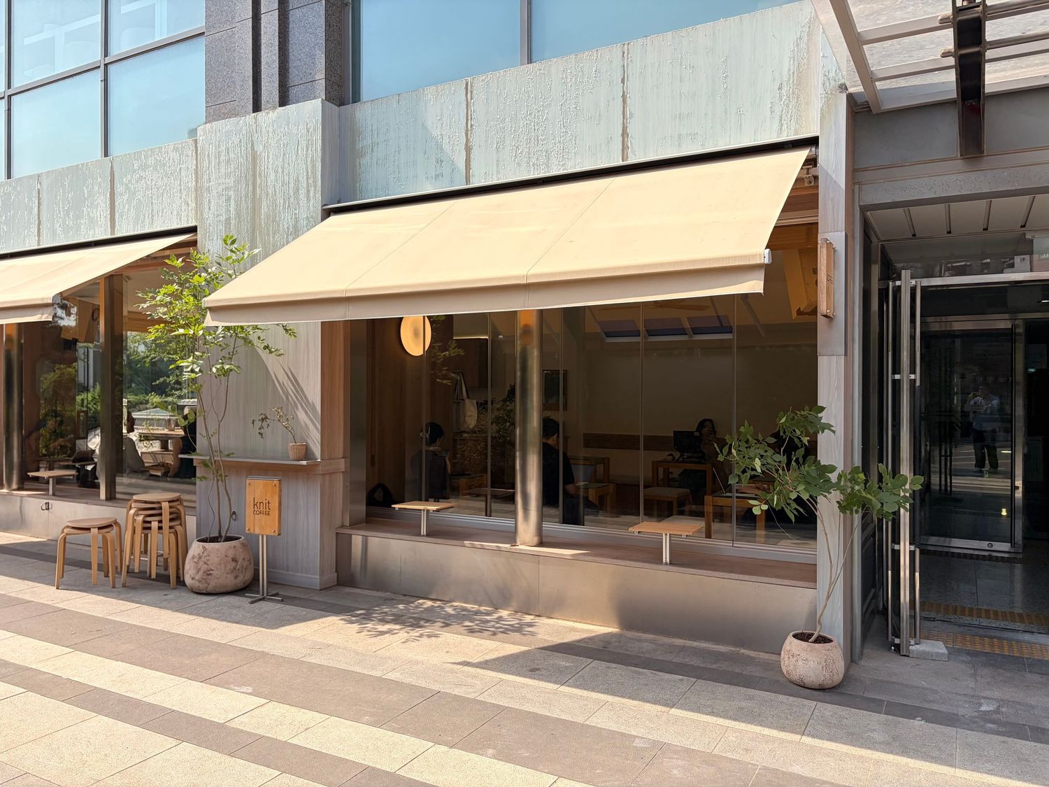 Modern cafe storefront with beige awning, large glass windows, and potted plants on a sunny urban sidewalk.