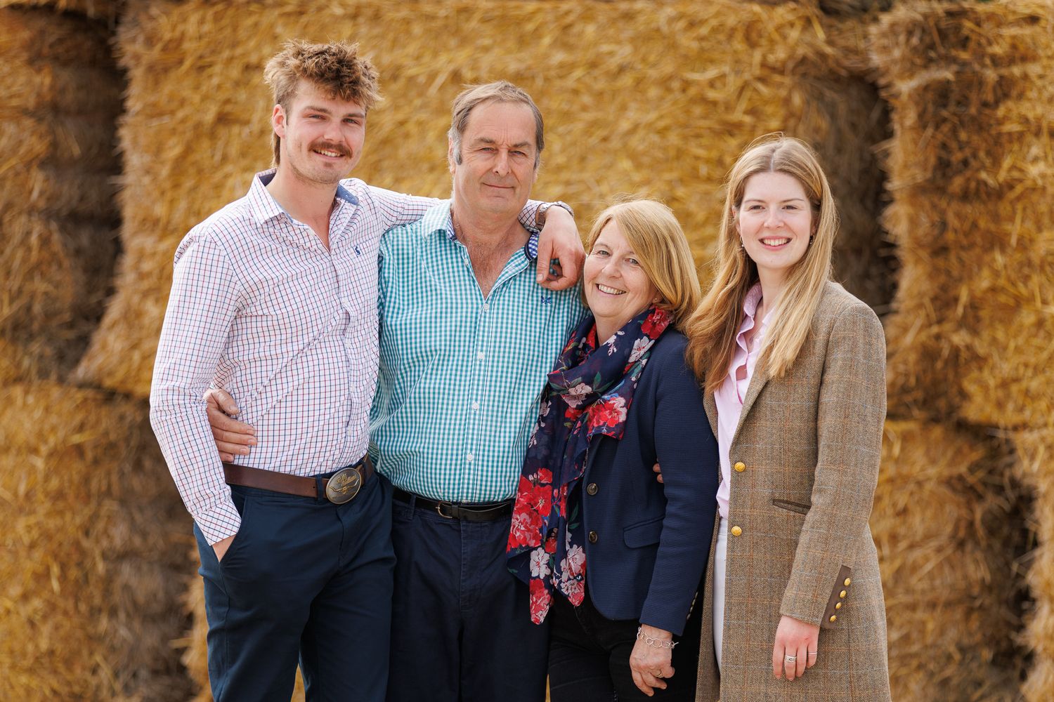 Family of four smiling together outdoors in front of hay bales on a farm.