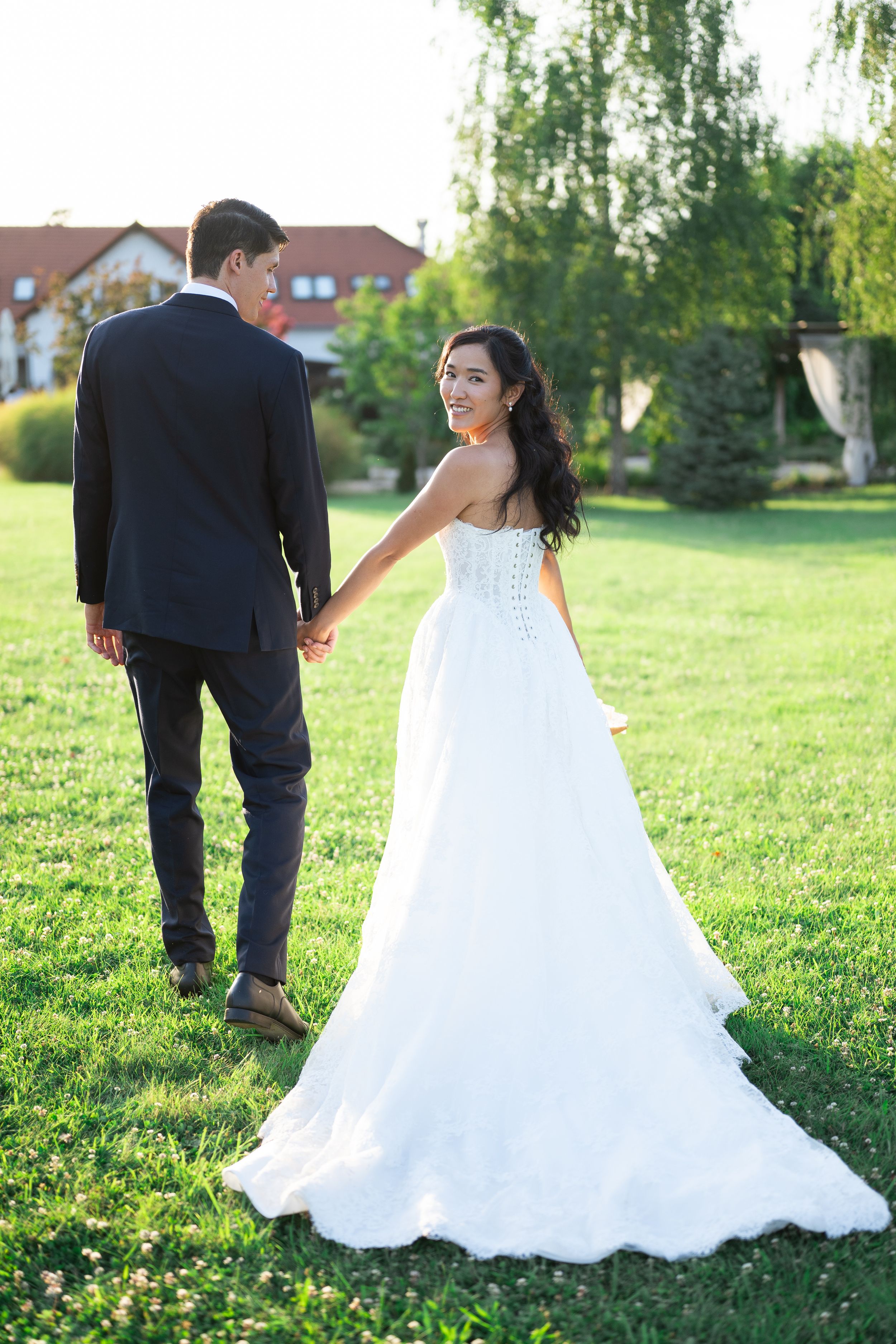 Smiling bride in white gown holds groom's hand walking on green lawn with trees and building in background.