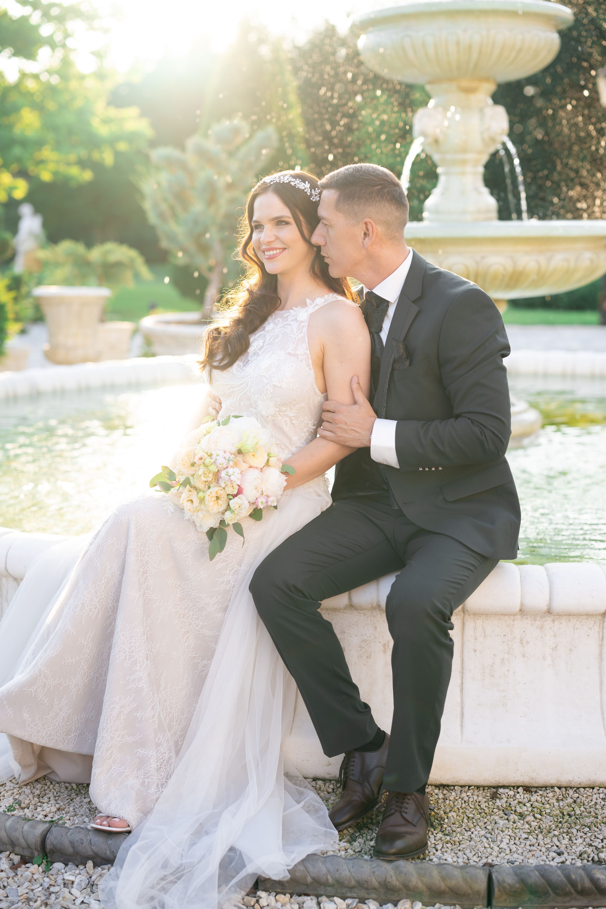 Bride and groom sitting by ornate stone fountain in sunlit garden, bride holding pink bouquet in lace wedding gown.