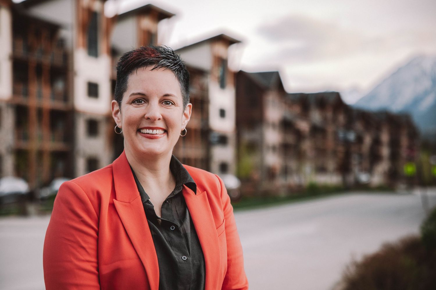 Professional woman in red blazer smiling outdoors with mountain backdrop and modern residential buildings.