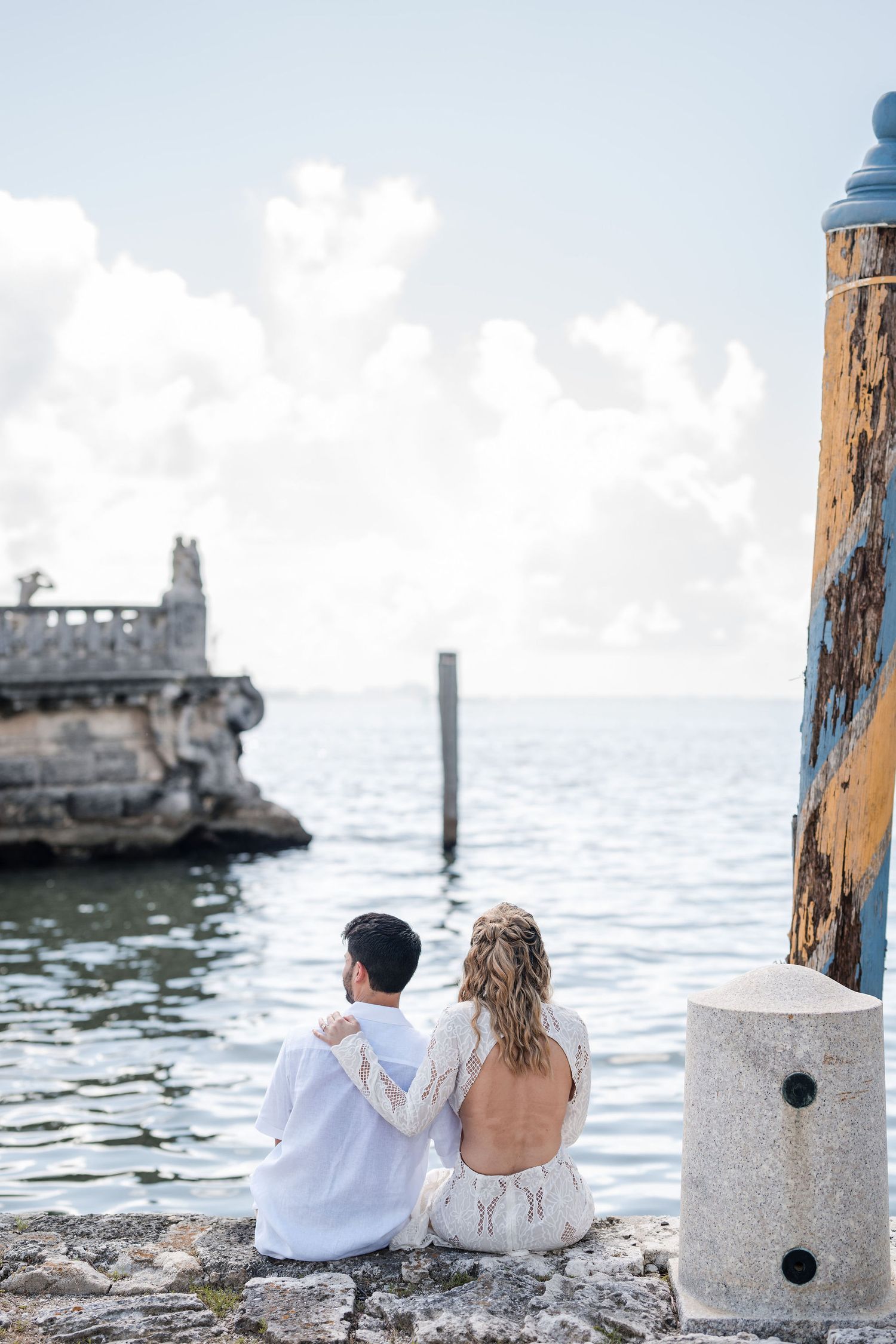 Bride and groom sit by the waterfront at Vizcaya Gardens, overlooking Biscayne Bay and the estate’s iconic stone barge.