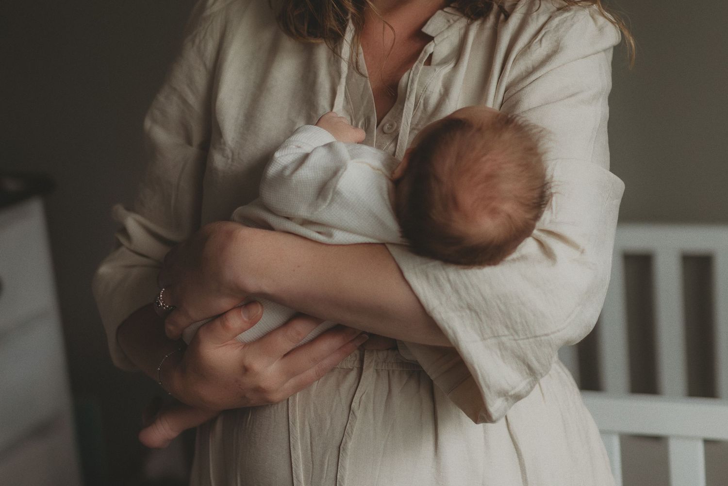Mother in linen dress gently cradling a newborn baby in her arms in a softly lit nursery room.