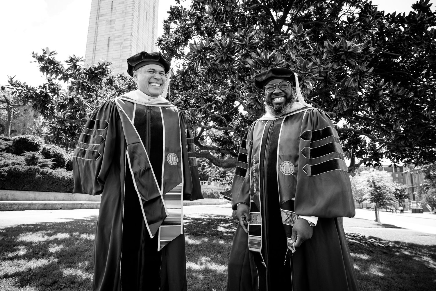 Two academics in doctoral regalia smile outdoors near trees and a tower at a university graduation ceremony.