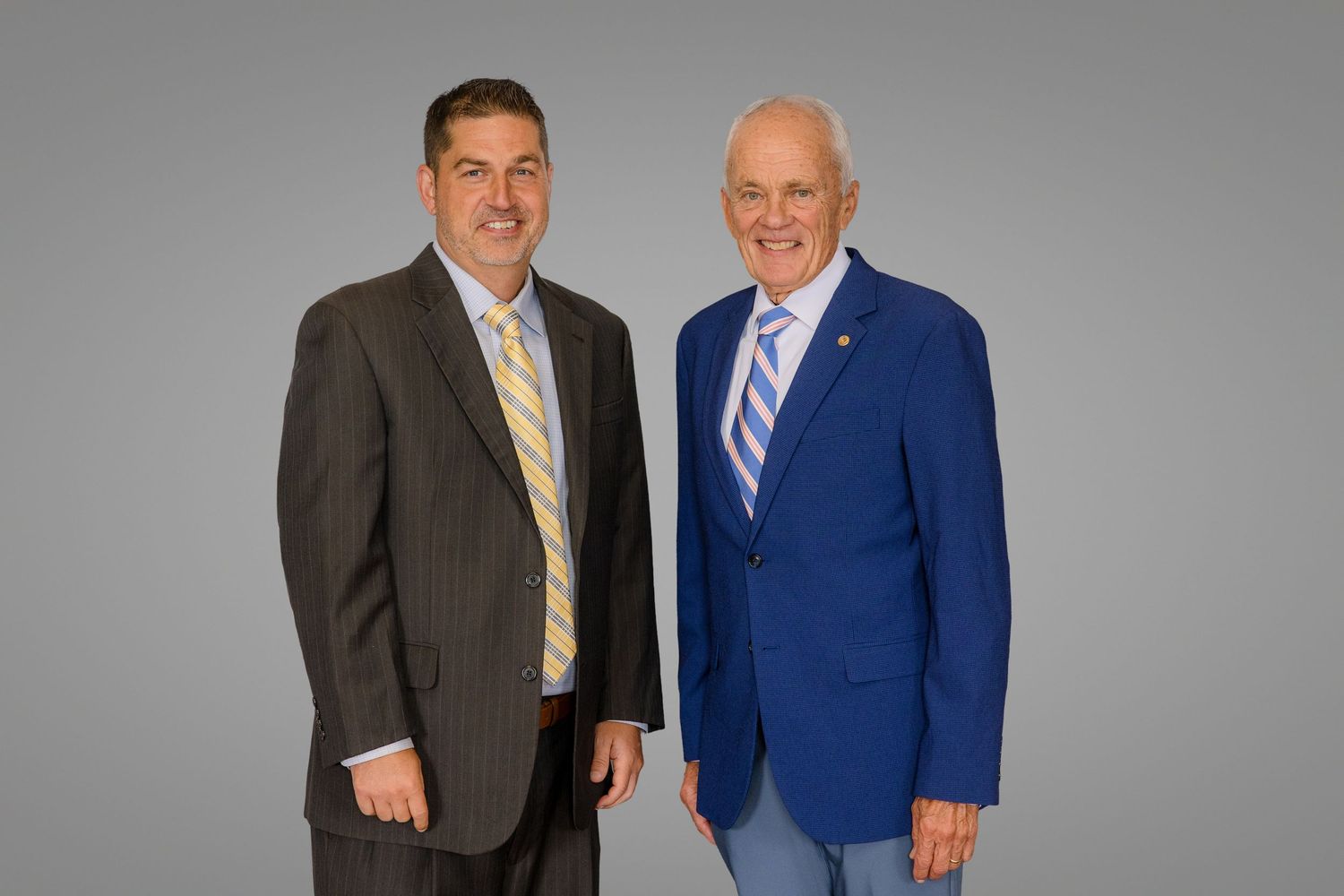 Two professional men in suits smiling, one in a blue blazer, posing together against a gray background.