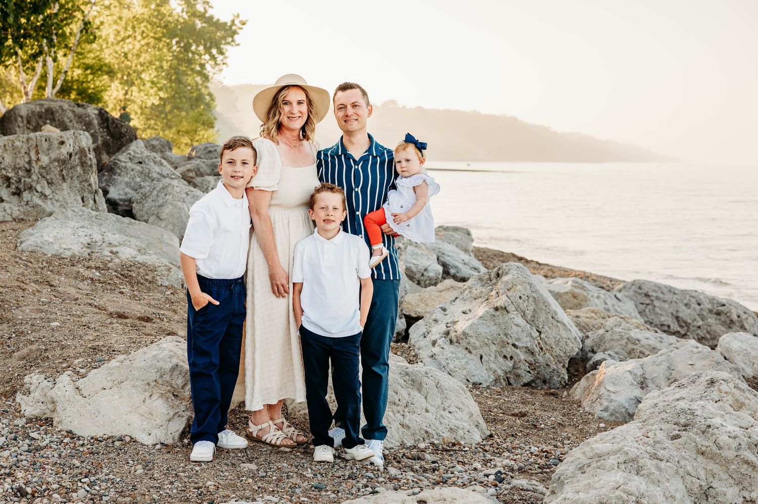 Family of five posing on rocky shoreline at sunset, parents with three children, wearing coordinated outfits in white and navy.