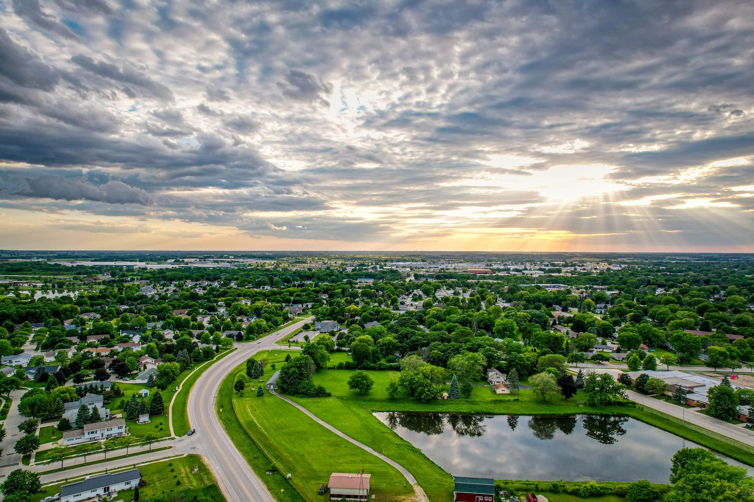 Aerial sunset view of a small Midwestern town with green parks, a pond, and dramatic golden rays breaking through clouds.