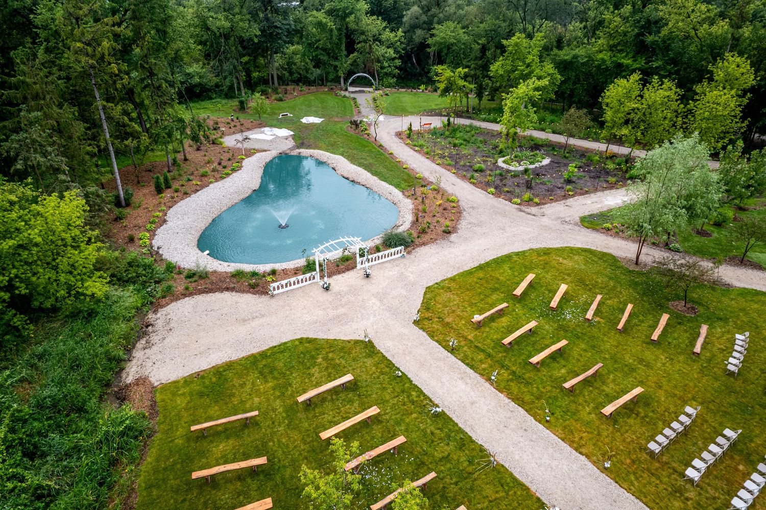 Aerial view of a lush garden with a kidney-shaped pool, pergola, and rows of wooden benches on manicured green grass.