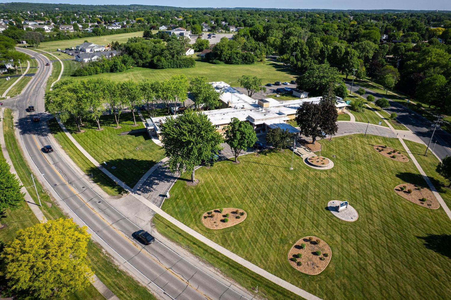Aerial view of a green park with circular garden beds along a busy suburban road.