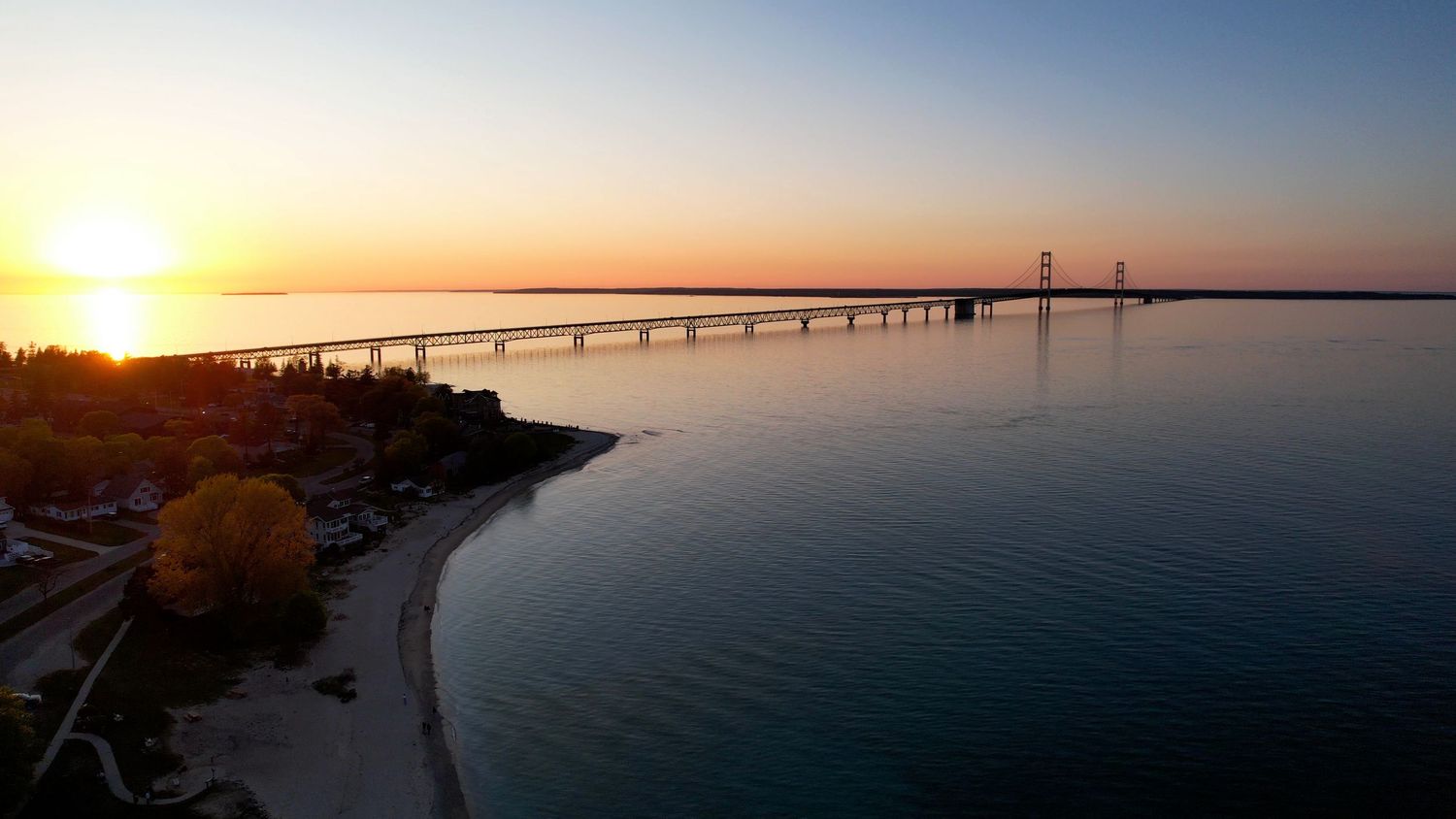 Aerial sunset view of the Mackinac Bridge spanning calm blue waters with vibrant orange and purple sky reflections.