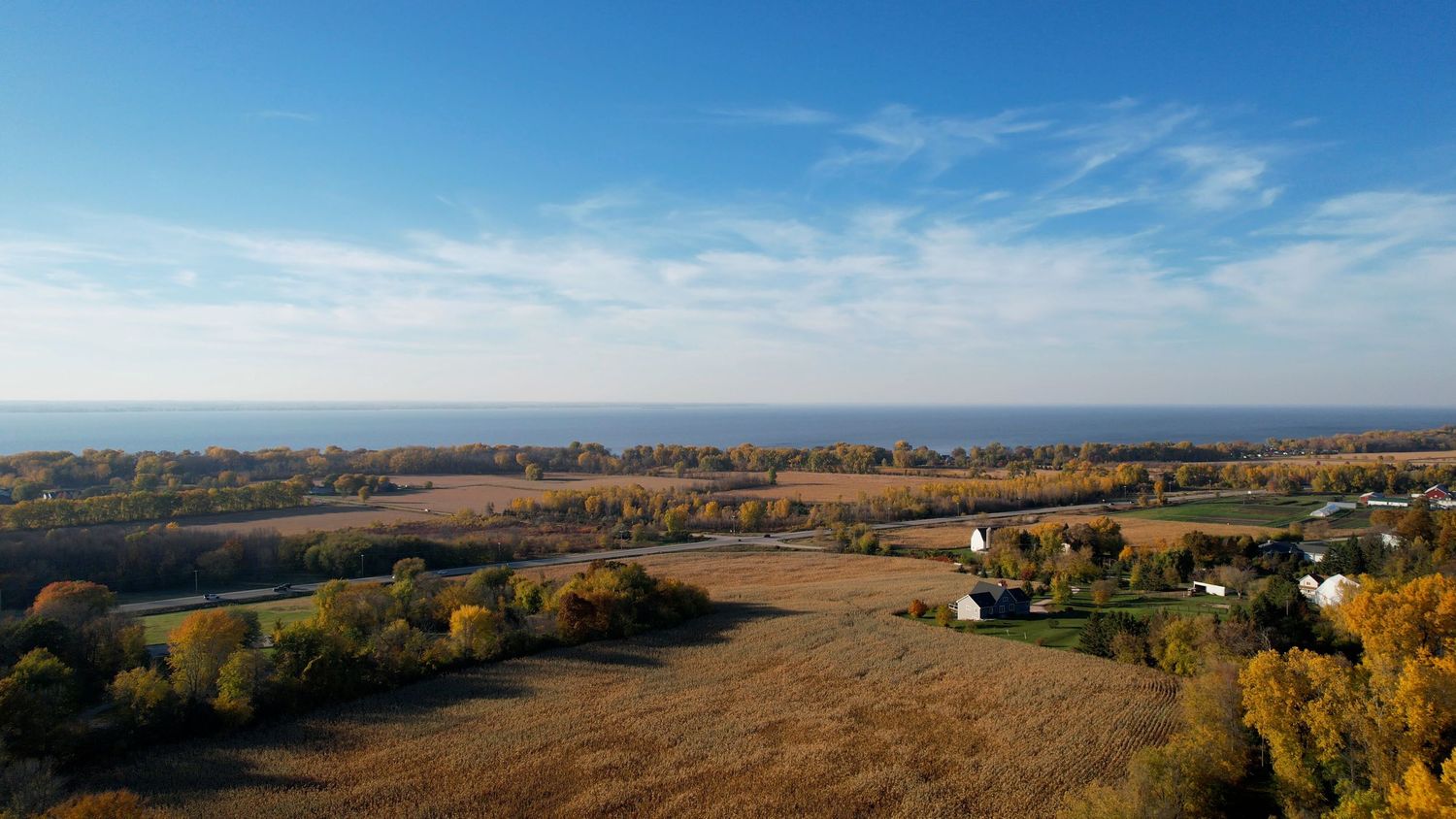 Aerial autumn view of flat farmland with harvested fields, colorful trees, and a lake visible on the horizon.
