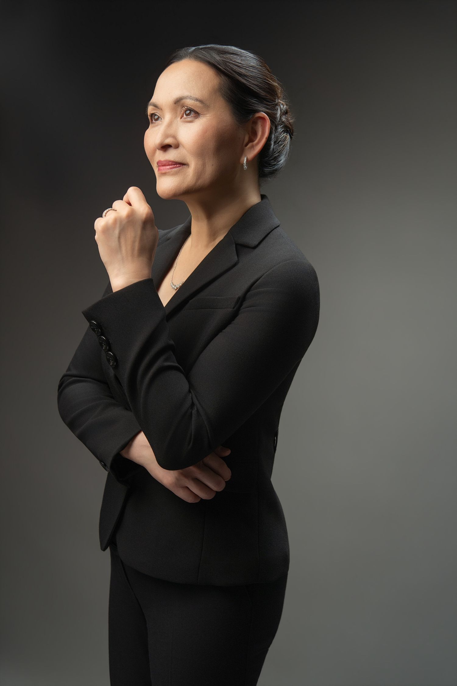 A confident businesswoman in a black suit poses thoughtfully with hand raised to chin against a dark gray background.