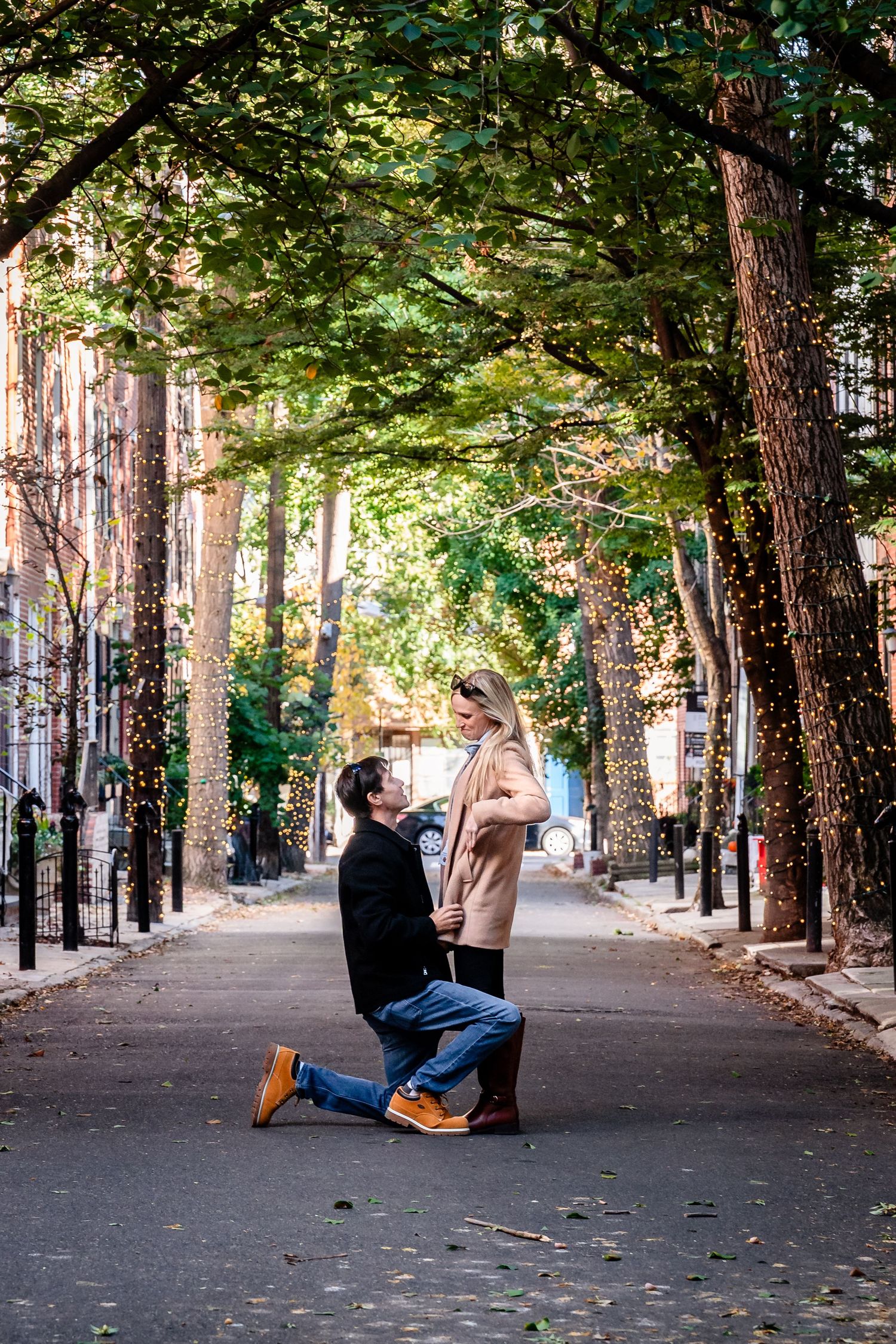Man proposing on one knee to woman on a tree-lined urban street decorated with fairy lights in autumn.