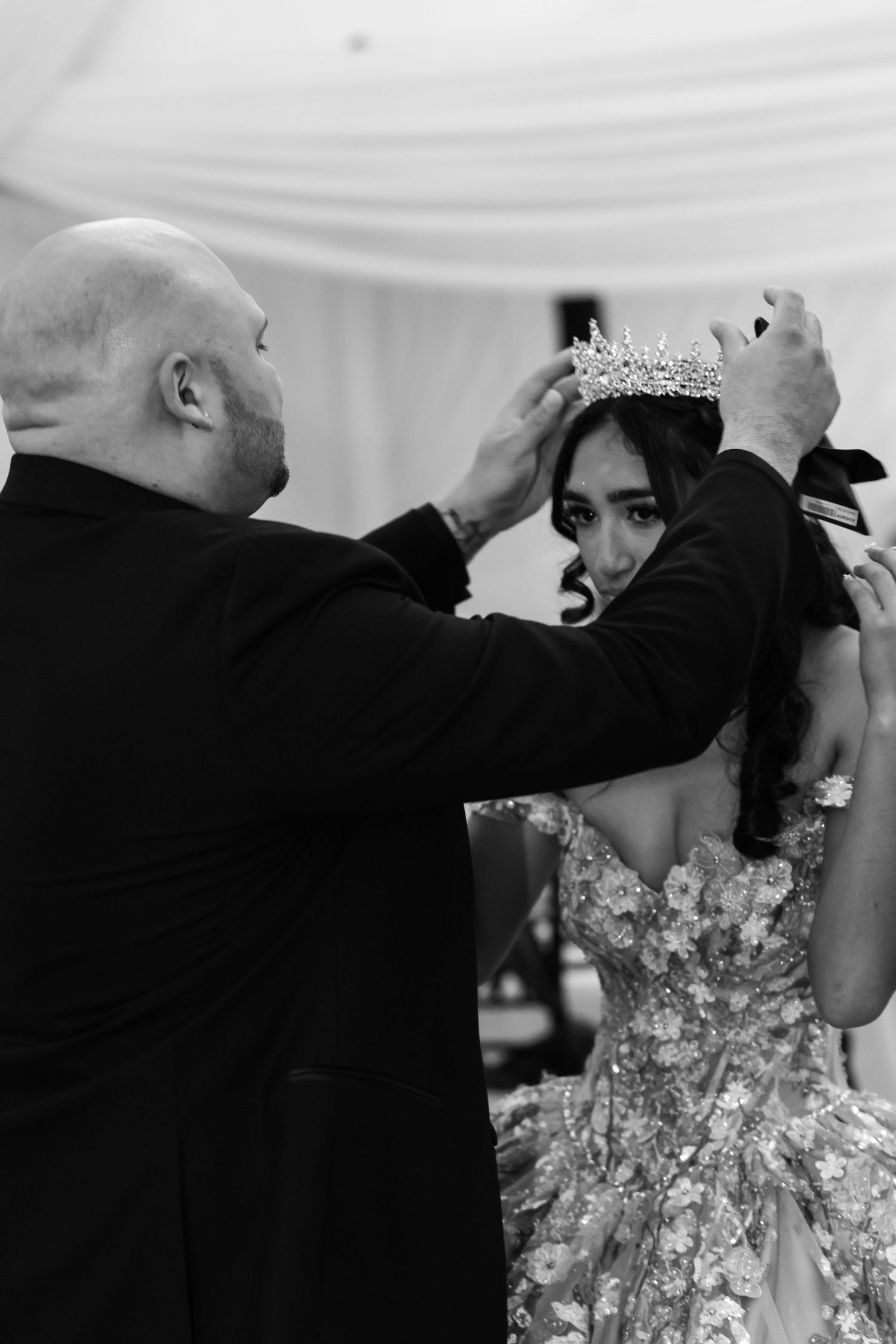 Man placing a crystal tiara crown on a bride in a floral wedding gown during a ceremony, black and white photo.