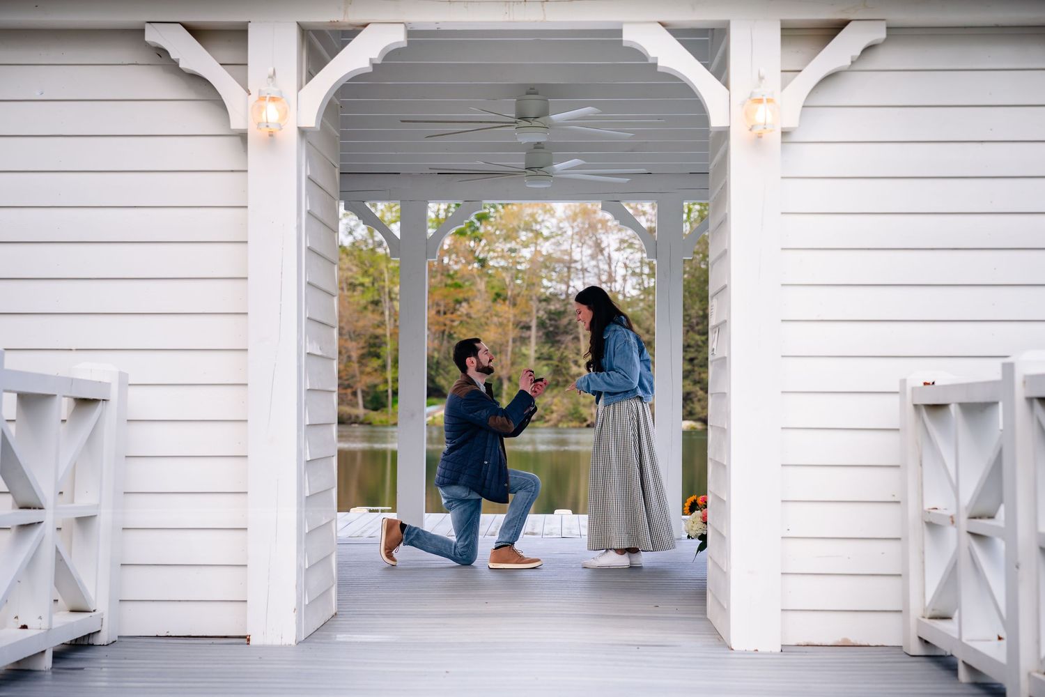 Man proposes on one knee to woman on white porch overlooking lake, romantic outdoor marriage proposal scene.