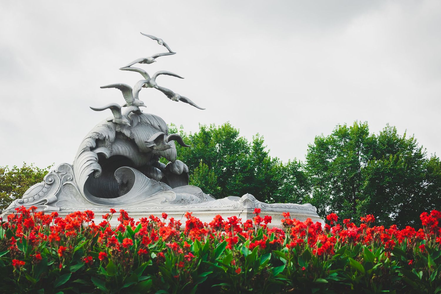 Navy merchant marine memorial stone sculpture of sea creatures and birds in flight, surrounded by vibrant red flowers and lush green trees.