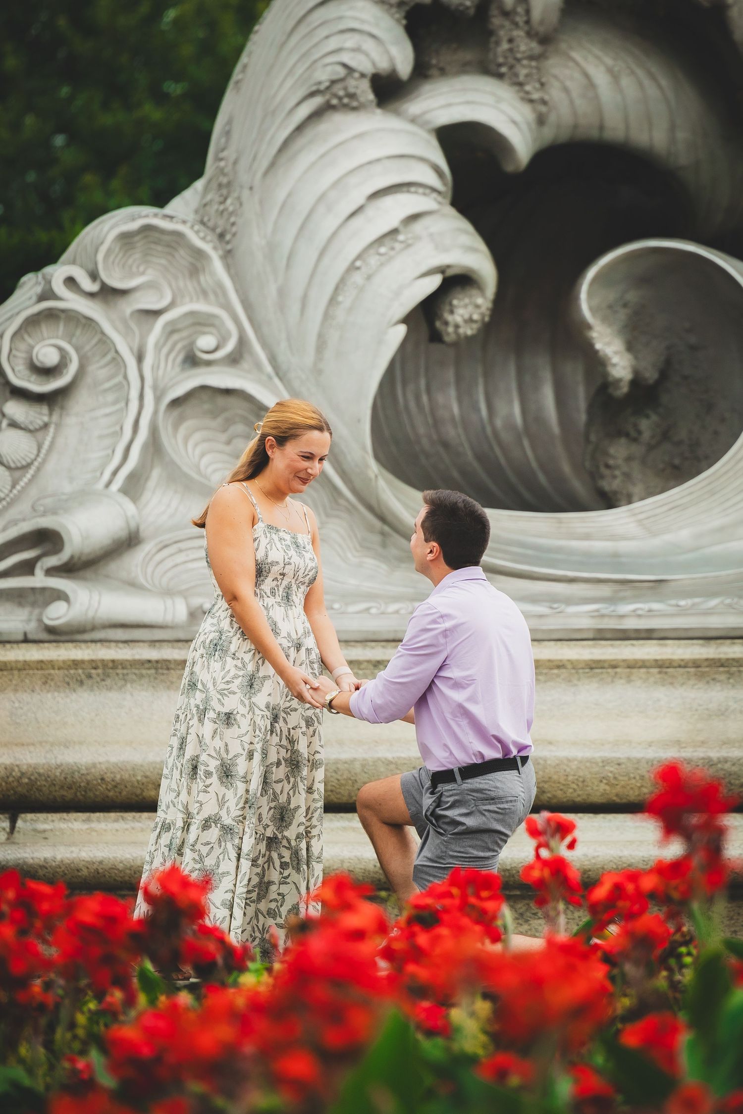 Man proposing on one knee to woman in floral dress by ornate stone fountain with red flowers.