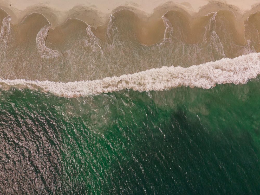 Aerial drone view of turquoise ocean waves crashing onto a sandy beach shoreline.