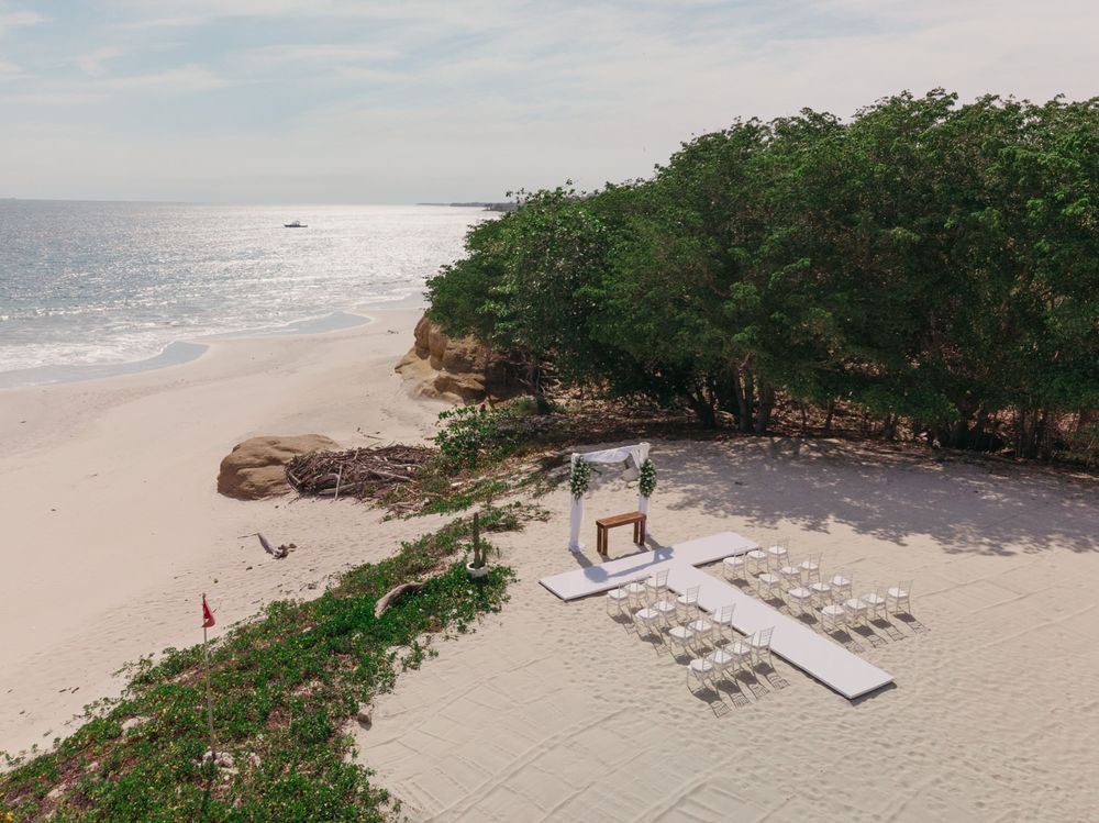 Aerial view of an elegant beach wedding setup with white chairs and floral arch beside lush green trees on a pristine sandy shore.