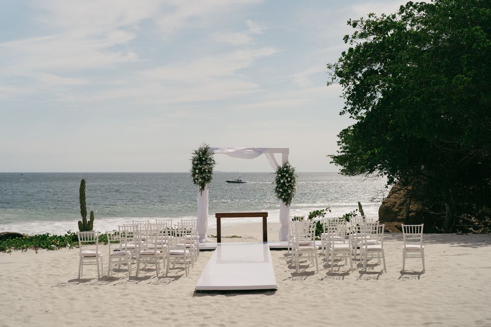 Elegant beach wedding ceremony setup with white arch, floral arrangements, and chairs on sandy shore.