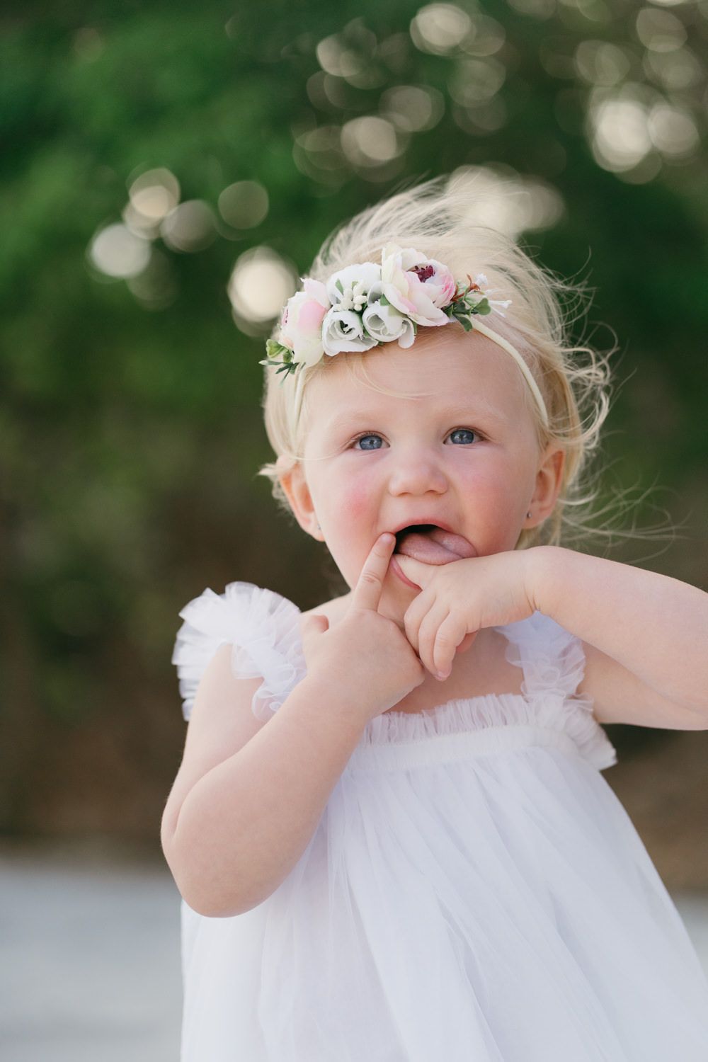 Adorable toddler girl in white dress and floral crown with finger in mouth, outdoors with green bokeh background.