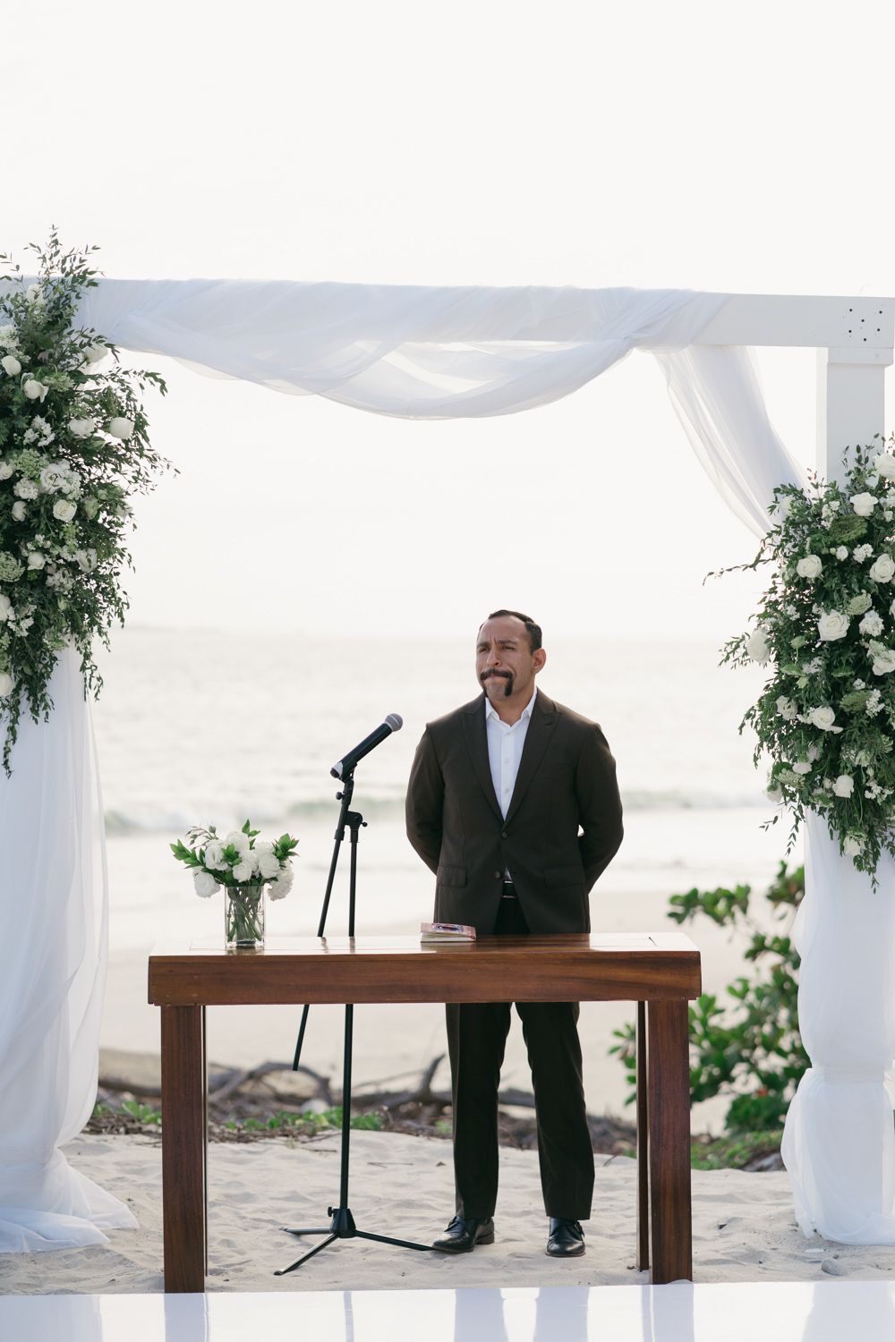 Officiant standing at wooden altar under white draped arch with white floral arrangements at beach wedding.