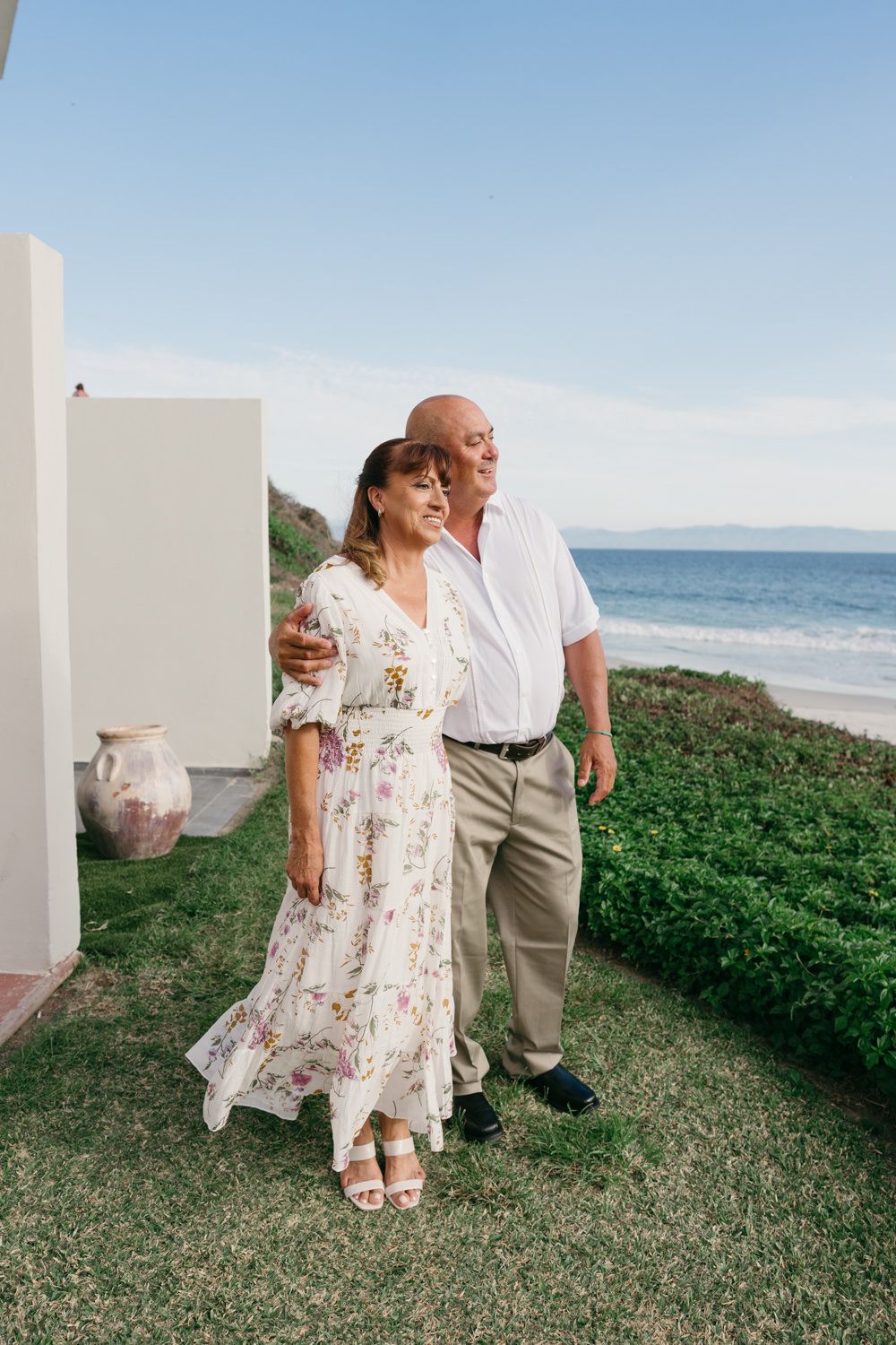 Happy older couple walking together in garden with ocean view, woman in floral dress, man in white shirt.