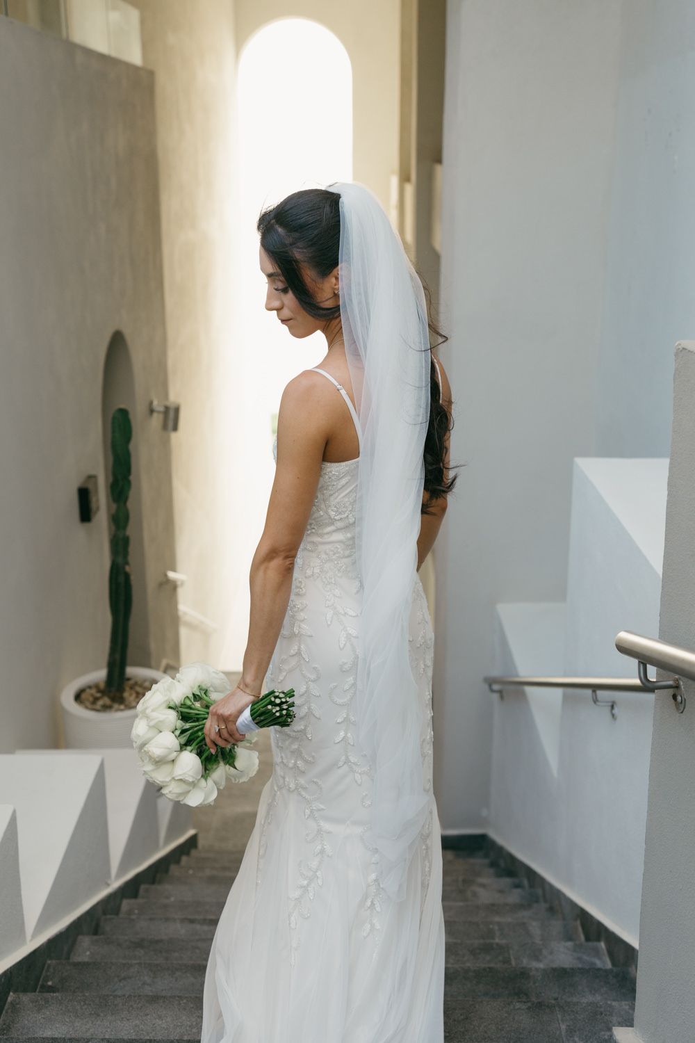 Bride in white lace gown and veil holds bouquet of white roses on staircase with cactus in background.