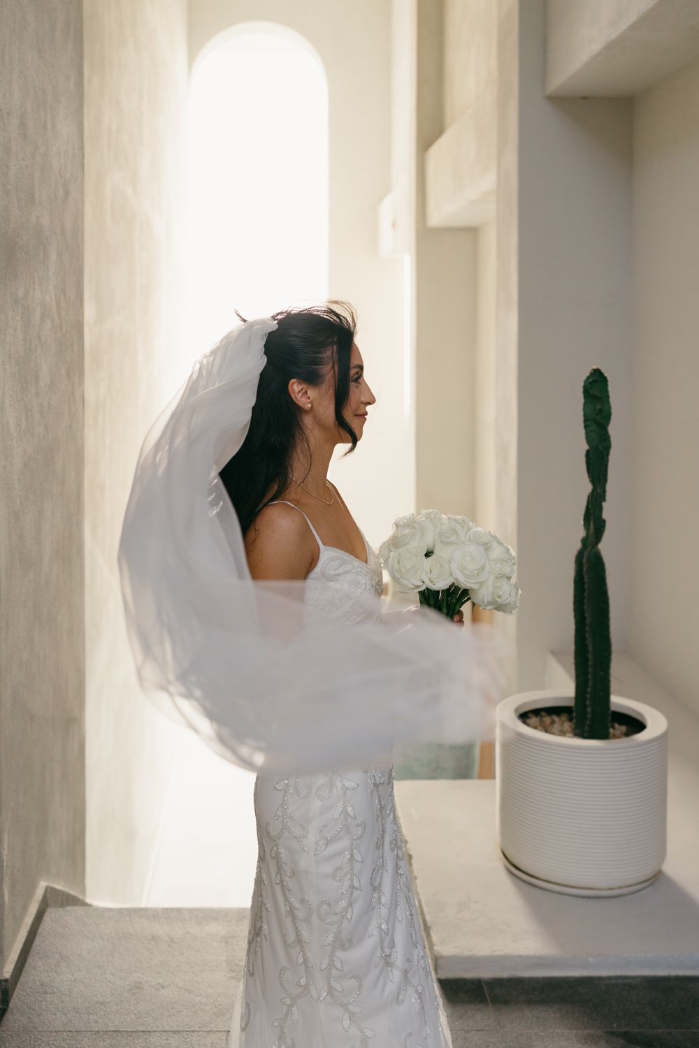 Bride in white lace gown and flowing veil holds bouquet of white roses, standing in sunlit archway near cactus.