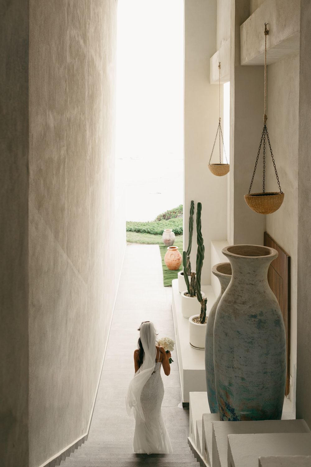 Bride in white gown holding bouquet walks through minimalist corridor with ceramic vases and cacti toward ocean view.