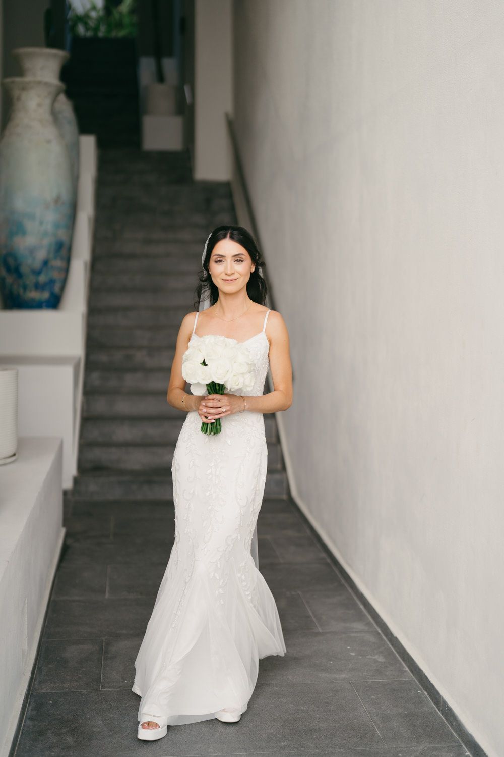 Elegant bride in fitted lace mermaid gown holds white bouquet while descending staircase in modern villa.